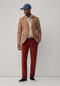 Blazer de tweed marrón, suéter beige con cremallera, pantalones de pana rojos, gorra azul y zapatillas negras. El atuendo presenta texturas suaves y colores contrastantes.