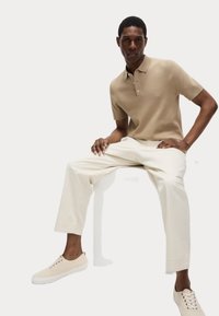 Polo de manga corta beige, corte ajustado, combinado con pantalones blancos de corte entallado y zapatillas beige. Textura suave, detalle de botón simple en el cuello.