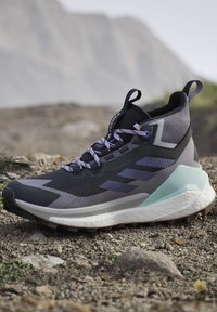 Chaussure de randonnée montante noire et grise avec des lacets violets et une semelle blanche épaisse sur un sol rocheux en extérieur.