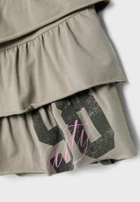Tissu vert olive clair avec volants superposés et imprimé du numéro 90 en noir, partiellement recouvert par le texte cursif rose « vanity ».