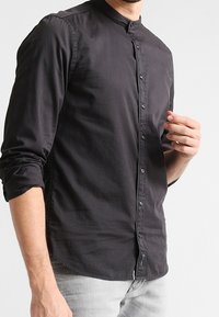 Camicia nera a maniche lunghe con colletto a banda e apertura frontale. Tessuto liscio e leggero con una leggera lucentezza.