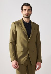Giacca su misura verde oliva con due tasche frontali, chiusura a un bottone e rever affilati. Indossata su una maglietta polo nera.