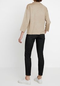 Person klädd i en beige långärmad topp, svarta slim-fit jeans och vita sneakers som står mot en vit bakgrund, vänd bort.
