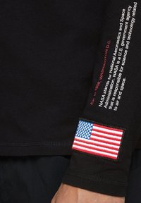 Camisa de manga larga negra con detalles en texto rojo que indican la fundación de la NASA y un parche de la bandera americana en la manga. Material de algodón, corte holgado.