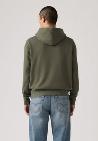 Κουκούλα πράσινου ελαιόχρου με κορδόνι στη κουκούλα, ριμπ μανσέτες και τελείωμα, συνδυασμένη με ανοιχτό μπλε τζιν τζιν με πίσω τσέπες και ξεθωριασμένη υφή.