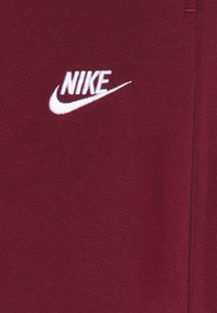 Vitt Nike-logotyp broderad på maroontyg med synlig söm på högersidan.