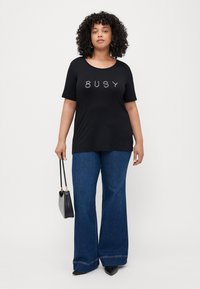 Svart T-skjorte med "BUSY" i hvit skrift, kombinert med vide blå denimjeans. Modellen holder en liten svart håndveske. Glatt tekstur.
