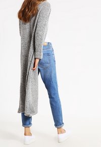 Lång, öppen cardigan i strukturerad grå garn, ihop med ljusblå slitna jeans och vita sneakers, sedd bakifrån.