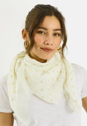 Mujer con cabello castaño recogido, que lleva pendientes de aro dorados y una bufanda blanca con pequeños puntos dorados sobre una camiseta blanca.