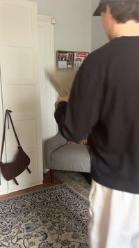 Osoba trzymająca beżową torbę stoi obok szarego fotela w pokoju z białą szafą i wzorzystym dywanem.