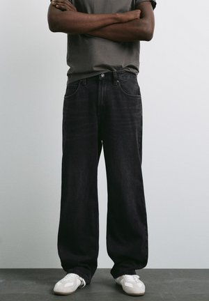 Mann trägt lockere schwarze Jeans, graues T-Shirt, weiße Sneaker und steht mit verschränkten Armen vor schlichtem hellem Hintergrund.