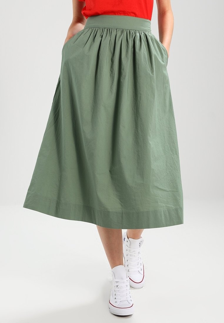 Femme portant une jupe verte mi-longue trapèze avec des poches et des baskets blanches montantes, assortie à un haut rouge.