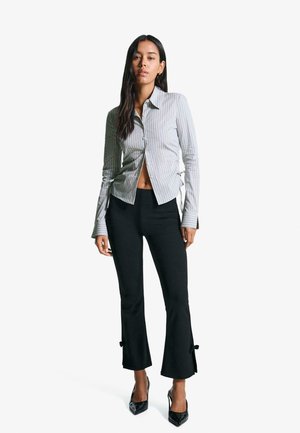 Gestreiftes Hemd mit cropped Design und seitlichen Bindebändern, kombiniert mit schwarzen, ausgestellt geschnittenen Hosen, die am Saum Schleifen haben, und schwarzen High Heels.