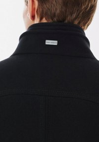 Czarna odzież wierzchnia z wysokim kołnierzem, z metalową metką z logo z tyłu. Materiał wydaje się być teksturowany z widocznymi detalami szycia.