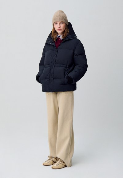 Giacca a vento nera con colletto alto, bottoni frontali e tasche laterali. Indossata sopra un maglione bordeaux e pantaloni larghi di colore crema.