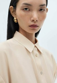 Bežová košeľa s golierom hladkej textúry a zapínaním na gombíky; model má na sebe zlaté kruhové náušnice, zelené oči a minimálny make-up.