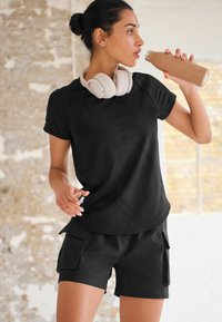 Sort kortærmet træningsskjorte parret med sorte cargo shorts. Har hvide over-ear høretelefoner og en beige vandflaske.