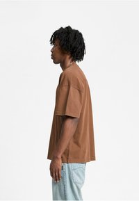 Brun, oversized t-shirt lavet af bomuld med korte ærmer, set fra siden. Har en afslappet pasform og minimale syninger.
