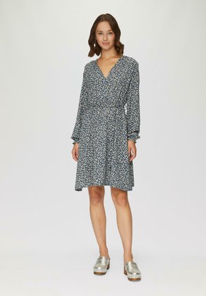 Robe fleurie bleue avec un décolleté en V, des manches longues et une ceinture à la taille. Présente une jupe fluide. Des sabots en argent complètent la tenue.