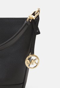 Schwarze Ledertasche mit strukturiertem Finish, einem goldenen MK-Logo-Anhänger und einem abnehmbaren Tragegurt mit einem polierten Goldverschluss.