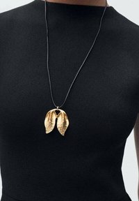 Collier en pendentif en forme de feuille d'or sur un cordon noir, présentant une surface texturée au design naturel et organique.