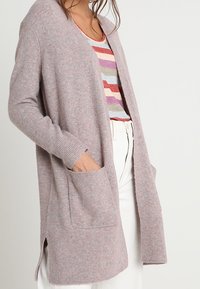 Cardigan rose clair avec un devant ouvert, des poignets côtelés et deux poches avant, fabriqué en matériau tricot doux. Fentes latérales à l'ourlet.