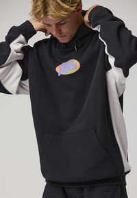 Felpa con cappuccio nera e grigia, dotata di tasca frontale, polsini a coste e logo iridescente. Il design include maniche a contrasto per un dettaglio in più.