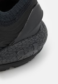 Acercamiento de un talón de zapatilla deportiva negra con tejido texturizado, suela de goma y un logotipo mate de Under Armour en el lateral.
