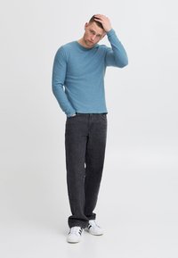 Chemise bleu clair à manches longues avec un col rond, associée à un jean gris foncé et des baskets blanches. Texture lisse, coupe décontractée.