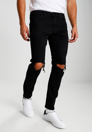Homme portant un jean skinny noir déchiré aux genoux, des baskets blanches, des chaussettes blanches et une chemise blanche sur un fond uni.
