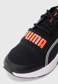 Pantofii atletici negri dispun de un guler din plasă, o brățară cu logo-ul îndrăzneț "PUMA" de culoare portocalie, talpă din cauciuc și detalii texturate.