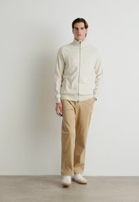 Ljust beige zip-up jacka i mjukt material, kombinerad med tanfärgade byxor och vita sneakers. Enkla, rena linjer med minimala designelement.