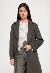Brown wool blend long coat with peak lapels, front pockets, and relaxed fit. Worn over a white top with feather-like texture and gray patterned pants.