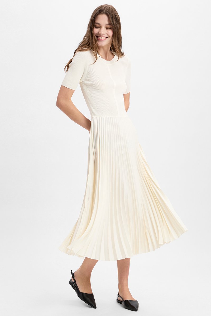 Cremefarbenes, plissiertes Midikleid mit kurzen Ärmeln, das über einen taillierten Oberteil und einen fließenden Rock verfügt. Kombiniert mit schwarzen Slingback-Pumps mit spitzer Spitze.