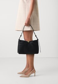 Borsa a tracolla in pelle nera con un design strutturato, dotata di una chiusura rotonda dorata e manico corto. Indossata con un vestito a quadri beige e décolleté color carne.