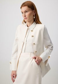 Kremowa, dopasowana kamizelka z złotymi guzikami, gładka w dotyku, z wysokim kołnierzem i długimi rękawami. Połączona z jasną koszulą i spodniami.