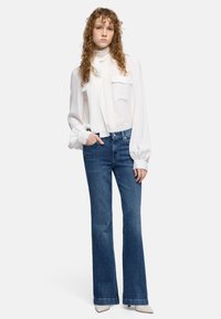 Jongere met krullend haar draagt een witte blouse met lange mouwen en strik bij de hals, en blauwe, hoge taille, flared jeans, staat met een hand in de zak.