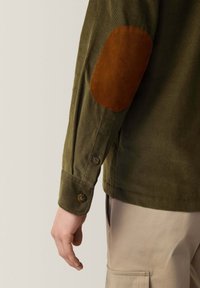 Camicia in corduroy oliva con toppe in suede marrone sui gomiti, polsini con bottoni e design aderente. Tessuto texture con righe visibili.