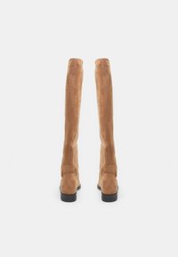 Botas de caña alta hechas de suave ante color beige, con un diseño ajustado, textura lisa y un tacón bajo negro.