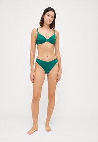 Τεχνητό μπικίνι σε τιρκουάζ απόχρωση με μπαλκονέ ψηλά με λεπτές τιράντες και αντίστοιχες χαμηλοκάβαλες βερμούδες, κατασκευασμένο από ομαλό, ελαστικό ύφασμα.