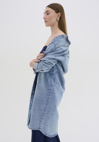 Abito camicia in denim azzurro chiaro con maniche lunghe, chiusura a bottoni e orlo curvo. La modella lo tiene sopra un vestito blu navy, mostrando look a strati.