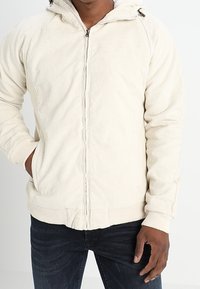 Man wearing a light beige corduroy zip-up jacket with hood and blue jeans, one hand in pocket, against a plain white background.
