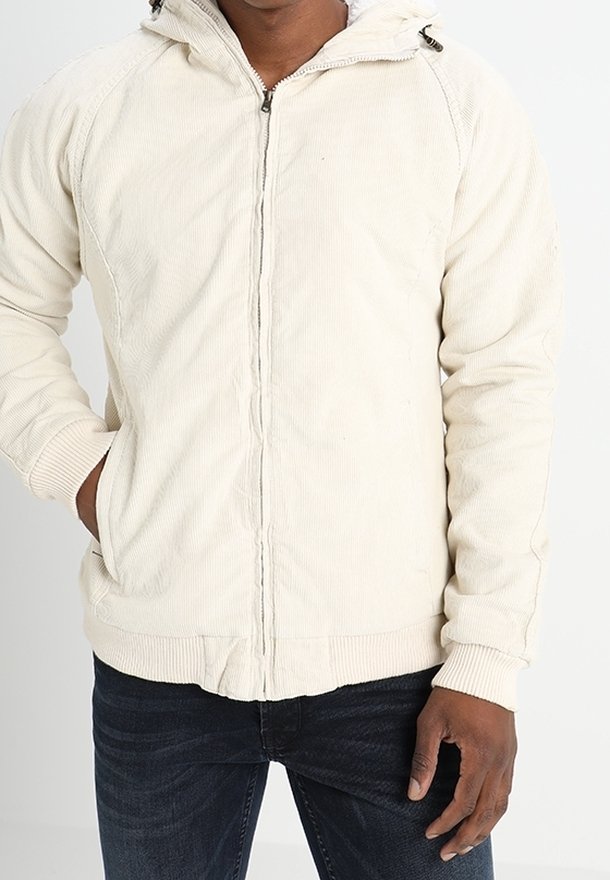 Man wearing a light beige corduroy zip-up jacket with hood and blue jeans, one hand in pocket, against a plain white background.