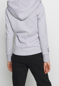 Sudadera gris con capucha, que presenta puños y dobladillo acanalados, textura suave y una pequeña etiqueta en la manga. Combinada con pantalones negros.