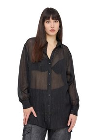 Camicia nera trasparente con finitura testurizzata, maniche lunghe e colletto, sovrapposta a un top a tinta unita. L'orlo cade sotto la vita.