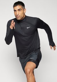 Uomo in abbigliamento sportivo Nike nero, in una posa di corsa a metà passo contro uno sfondo chiaro e semplice.