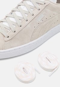 Béžová semišová teniska s bílou gumovou podrážkou, s šněrováním, perforovanými detaily a dvěma svazky bílých a béžových tkaniček.