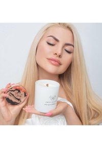 Biała świeca w cylindrycznym pojemniku z czarnym logo, drewnianym knotem i różowo-złotą pokrywką, trzymana przez osobę z długimi blond włosami.