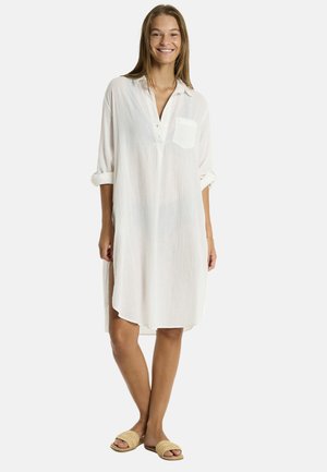 Donna sorridente che indossa un vestito bianco, con bottoni, lungo fino al ginocchio con maniche arrotolate e una tasca, abbinato a sandali casual, su uno sfondo semplice.