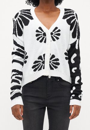 Femme portant un cardigan à boutons noir et blanc à motif floral et un jean noir, debout les bras détendus le long du corps.